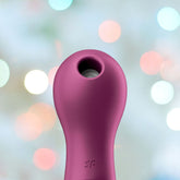 A Satisfyer Lucky Libra Air Pulse Clit Stimulator, featuring a pink, cylindrical shape with a rounded tip and circular opening, is set against a soft, blurred background with colorful bokeh lights, capturing the gentle allure of its design for clitoral stimulation.